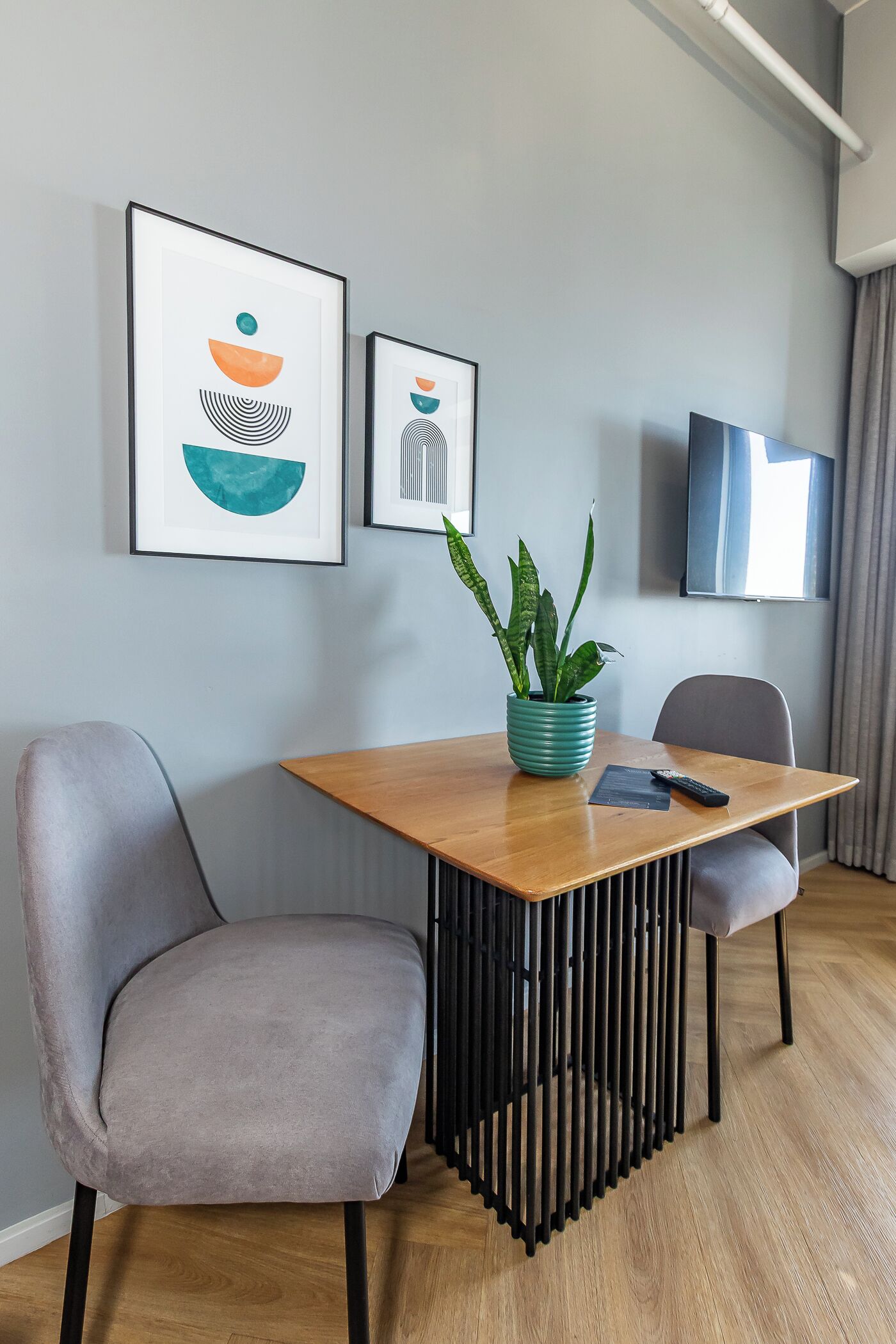 A modern room with a wooden dining table, two grey chairs, a potted plant, framed artwork, and a TV on the wall.