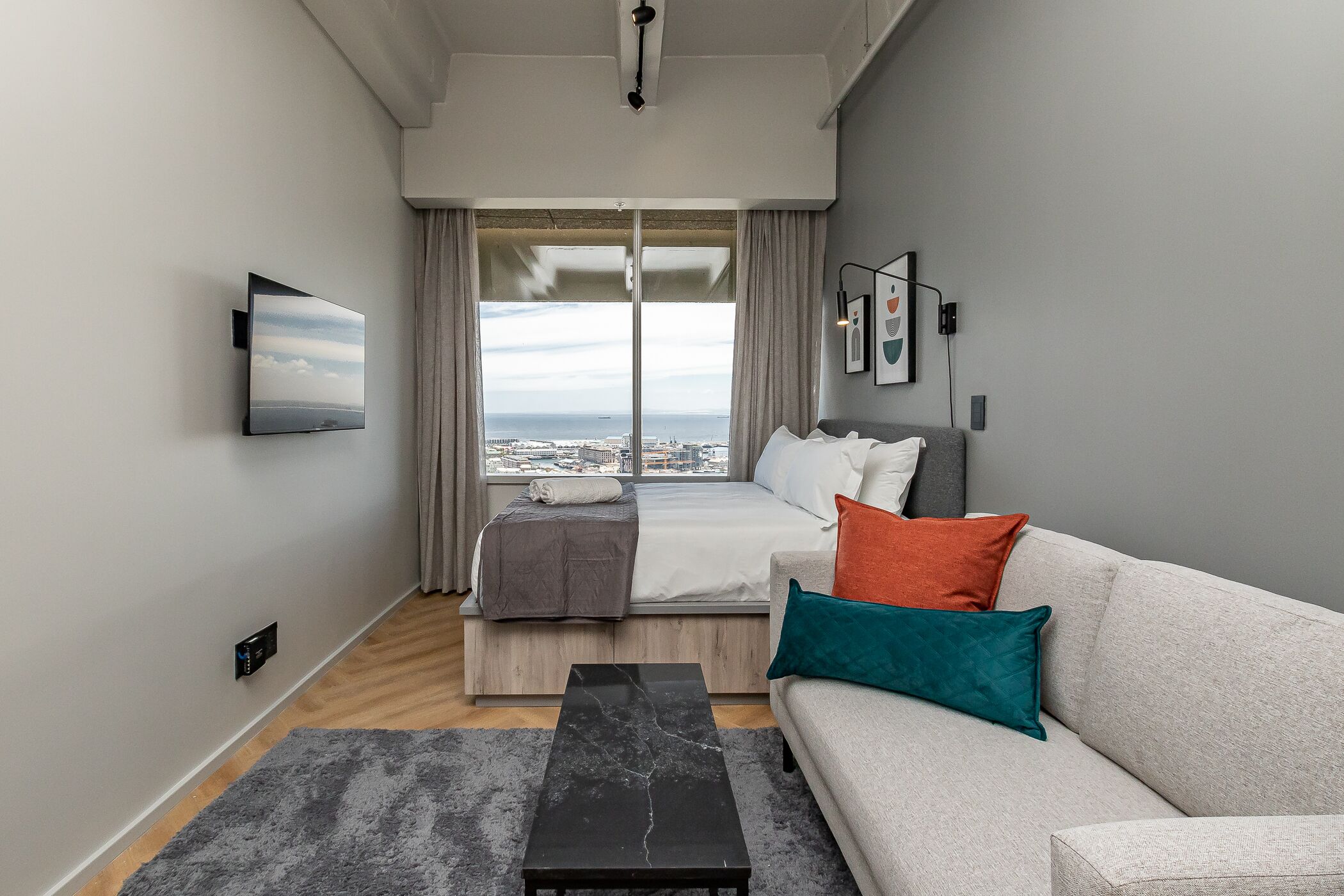 A modern hotel room with a bed, a sofa, a coffee table, and a large window offering a city view.