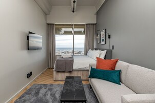 A modern hotel room with a bed, a sofa, a coffee table, and a large window offering a city view.