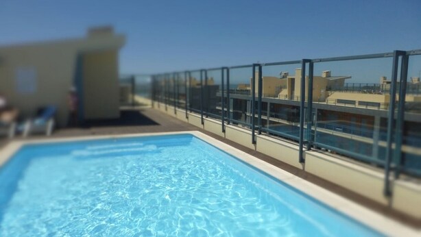 En swimmingpool med en glasbarriere, klar himmel og bygninger i baggrunden.