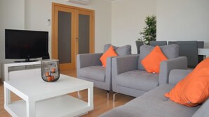 En stue med et fladskærms-tv, grå sofaer med orange puder, et hvidt sofabord og en potteplante.
