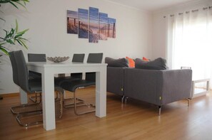 A modern living room with a white dining table, grey sofa, and a large window with curtains.