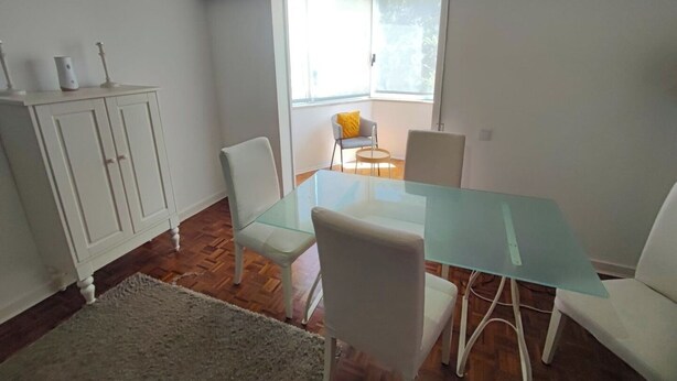 A dining area with a glass table, white chairs, a white cabinet, and a window with a view. - Carcavelos, Cozy 3 bedroom apartment in Cascais (Carcavelos)