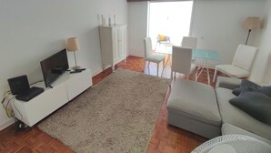 A living room with a sofa, a TV, a cabinet, a dining table with chairs, and a large window. - Carcavelos, Cozy 3 bedroom apartment in Cascais (Carcavelos)