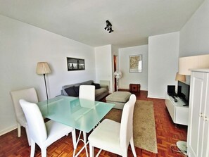 A modern living room with a glass dining table, white chairs, a sofa, a small side table, a fan, and a television.