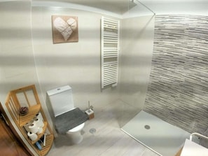 Une salle de bain Ă©quipĂ©e de toilettes, dâune douche et dâune Ă©tagĂšre en bois oĂč sont rangĂ©s les articles de toilette.