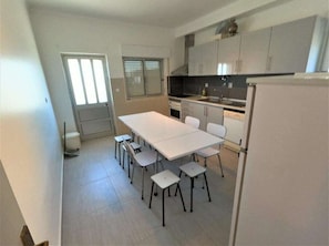 A kitchen with white cabinets, a white table, and chairs. There is a window with blinds, a door, and a refrigerator.