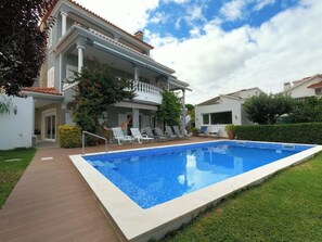A two-story house with a swimming pool, a covered patio with lounge chairs, and a well-maintained garden.
