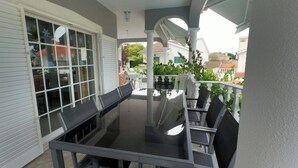 Un espace extérieur abrité, aménagé pour les repas, avec une table et des chaises en verre, une porte-fenêtre coulissante et une vue sur un quartier résidentiel.