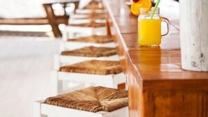 A row of white wooden chairs with woven cushions, a wooden bar with a glass of orange juice and a straw, and a white pillar in a covered outdoor area. - Akti Retzika (Thermaikos)