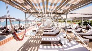 A beachside lounge area with wooden loungers and a hammock, shaded by striped canopies. - Akti Retzika (Thermaikos)
