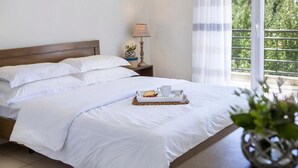 A neatly made bed with white linens, a tray with a cup and a pastry, a bedside lamp, and a view of greenery through a window. - Akti Retzika (Thermaikos)