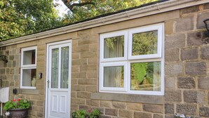 A stone building with a white door and four windows. There are two potted plants on the ground. - Bronte Cottage (Keighley)