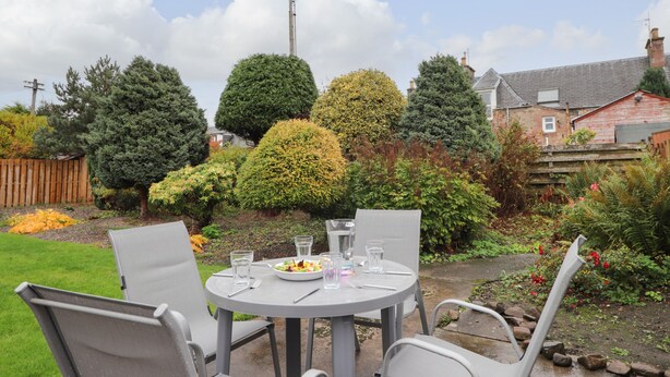 A round table with four chairs in a garden setting, surrounded by various trees and a wooden fence.