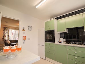 Une cuisine avec des meubles de couleur verte, un réfrigérateur blanc et un four noir. Il y a une horloge au mur et un saladier rempli de fruits sur le plan de travail.