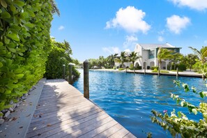 Una piscina con un muelle de madera, rodeada de exuberante vegetación y un edificio blanco con arcos al fondo.