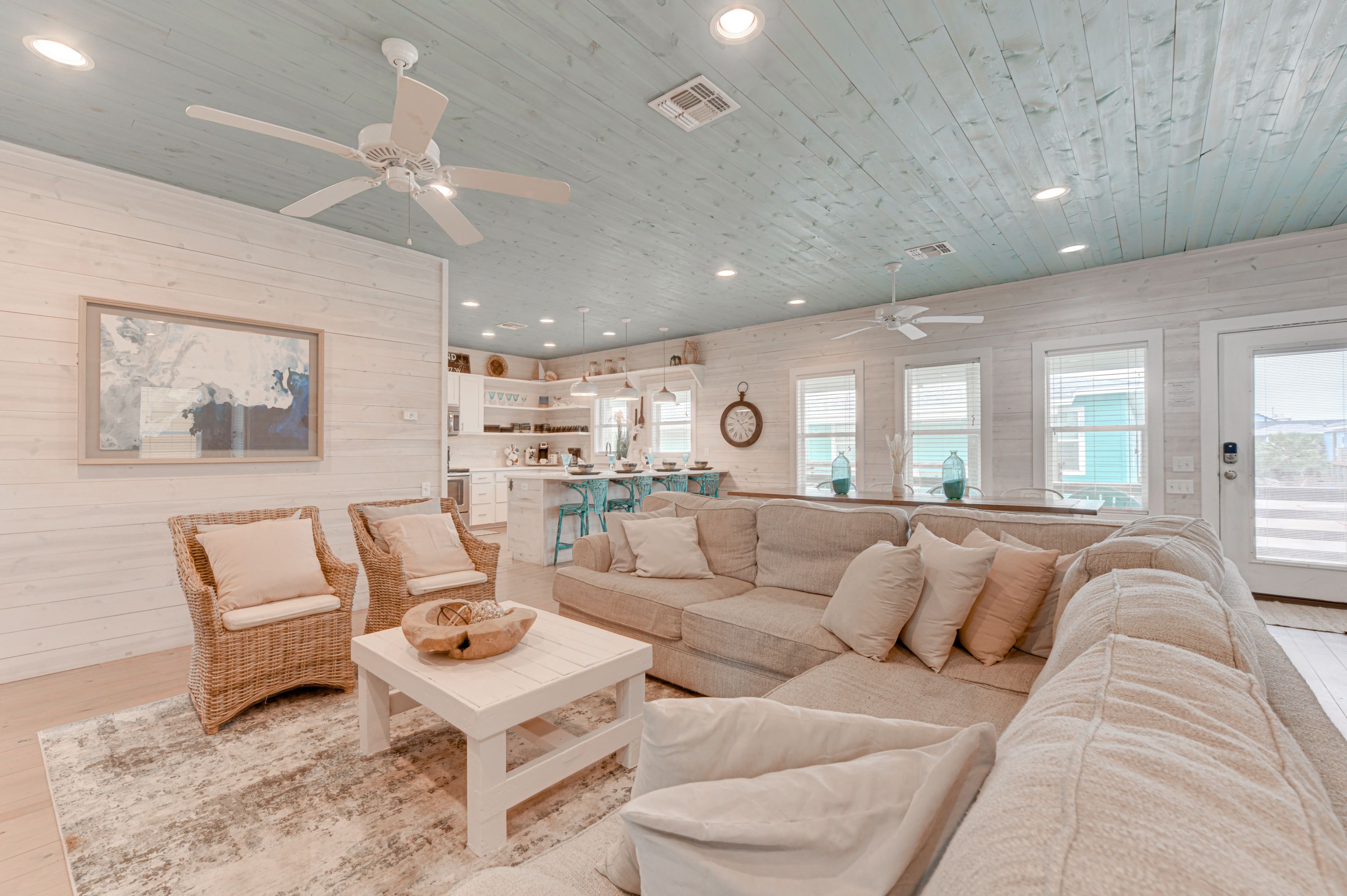 A spacious living room with a large beige sectional sofa, two wicker chairs, a white coffee table, and a ceiling fan.