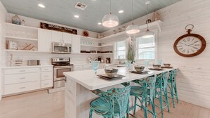 Une cuisine avec des meubles blancs, un îlot central, des chaises de couleur turquoise et une horloge murale.