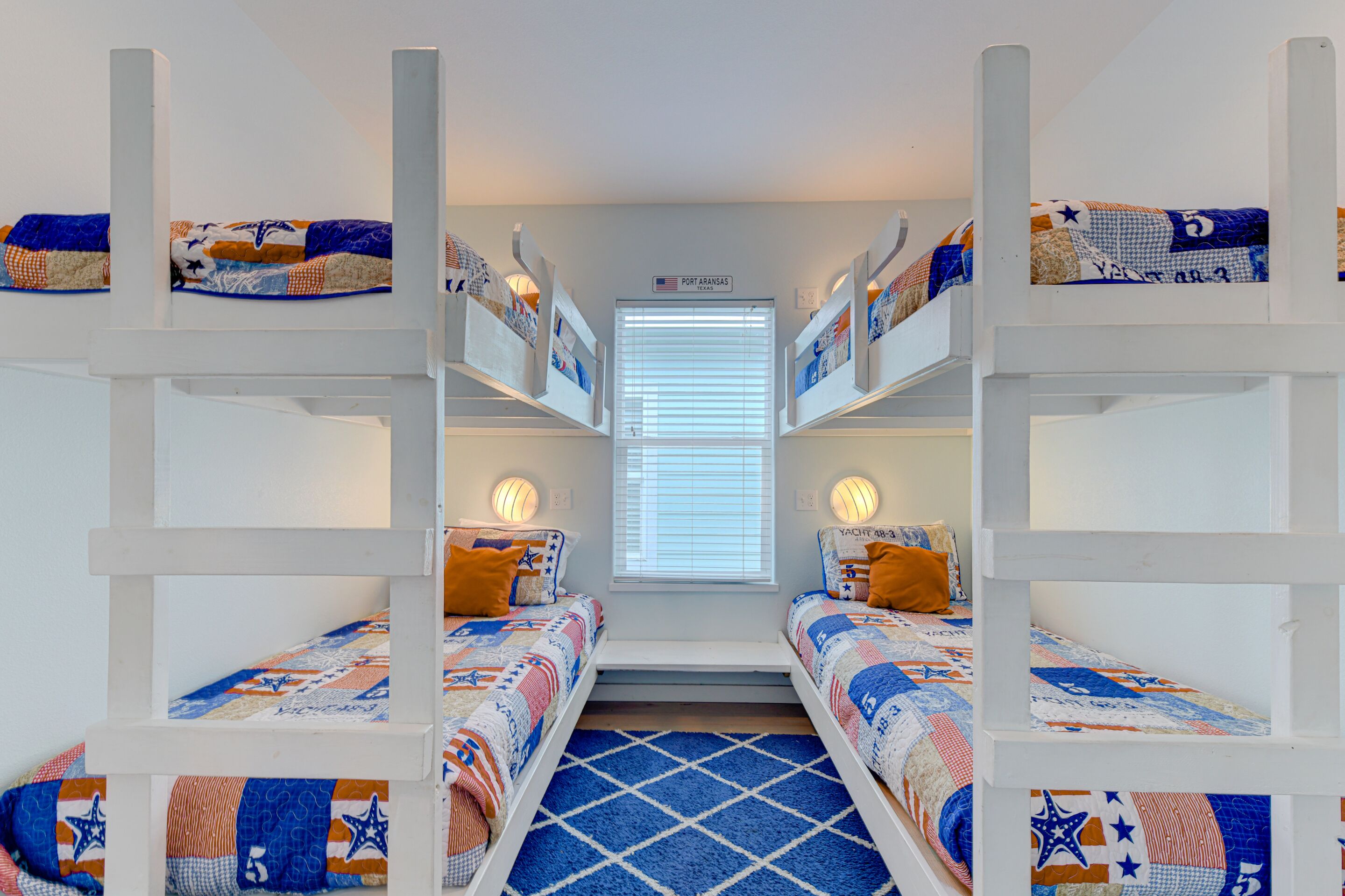 Une chambre avec deux lits superposés, chacun doté d’une tête de lit matelassée, d’une fenêtre avec des stores et d’une pancarte au-dessus de la fenêtre indiquant « Port Aransas ».