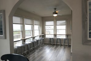 A room with a high ceiling, large windows, and a ceiling fan. There are multiple bar stools and a table.