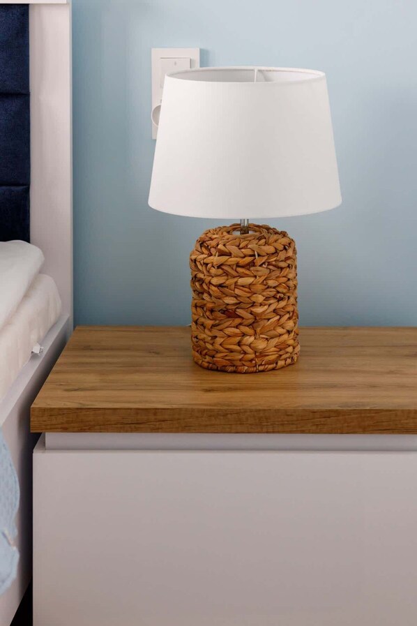 A lamp with a woven base on a wooden nightstand in a room with a blue wall and a white bed.