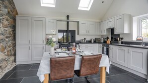 A modern kitchen with white cabinets, a dining table set for two, and a stone wall on one side. - Hen Felin (Pwllheli)