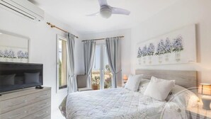 Ein Schlafzimmer mit einem Bett, einem Fernseher, einem Schminktisch, einem Deckenventilator und einem Fenster mit Vorhängen.