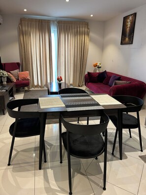A modern living room with a dining area, featuring a red sofa, a black dining table with chairs, and a large window with curtains. - Lennox 1bed (Accra)