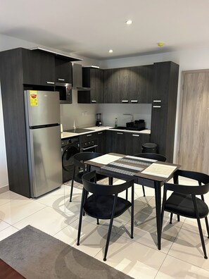 Une cuisine moderne avec des meubles de couleur sombre, un réfrigérateur en acier inoxydable, un four encastré et un coin repas comprenant une table et des chaises.