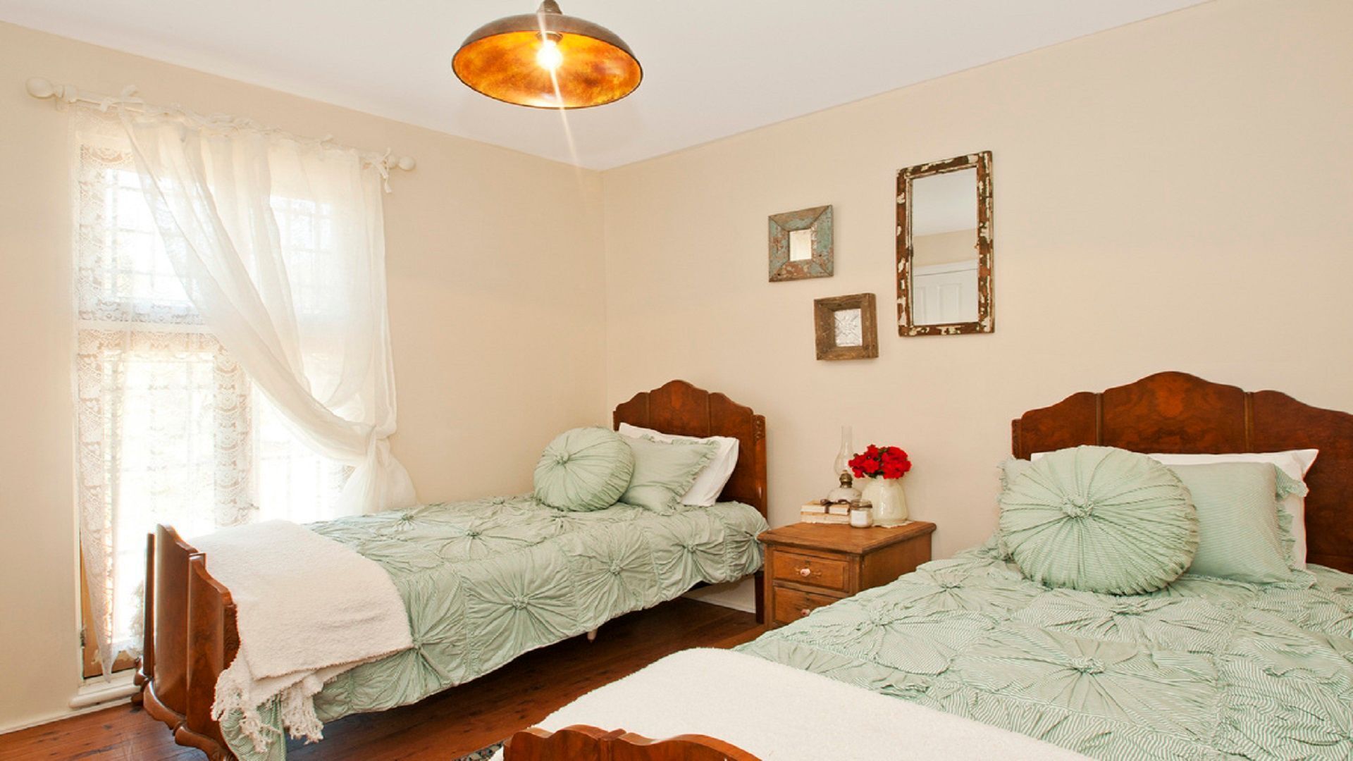 A bedroom with two single beds, a wooden nightstand with a vase of red flowers, and a mirror on the wall.