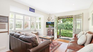 A cozy living room with a brown leather sofa, a coffee table, and a TV. There are large windows with blinds, a sign reading "RUE DE LA MARTILLIERE," and a view of greenery outside.