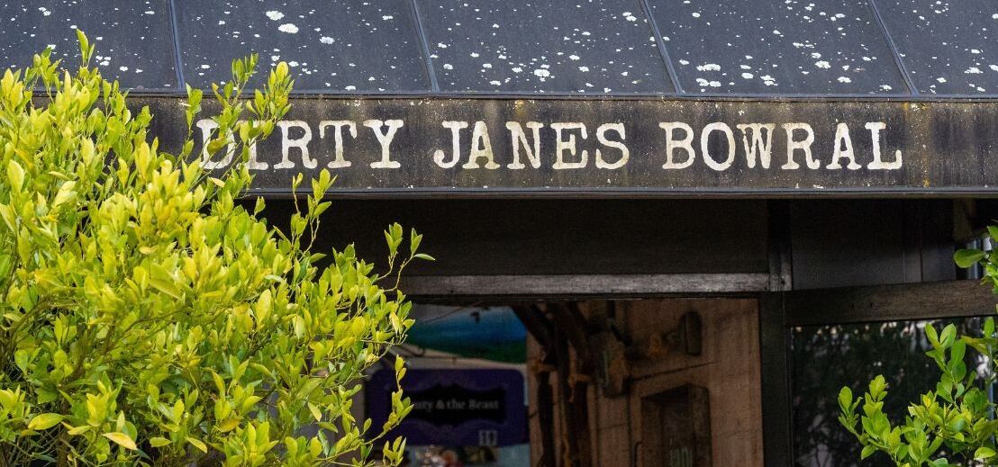 A sign for "Dirty Jane's Bowral" with a rustic wooden structure and greenery in the foreground.