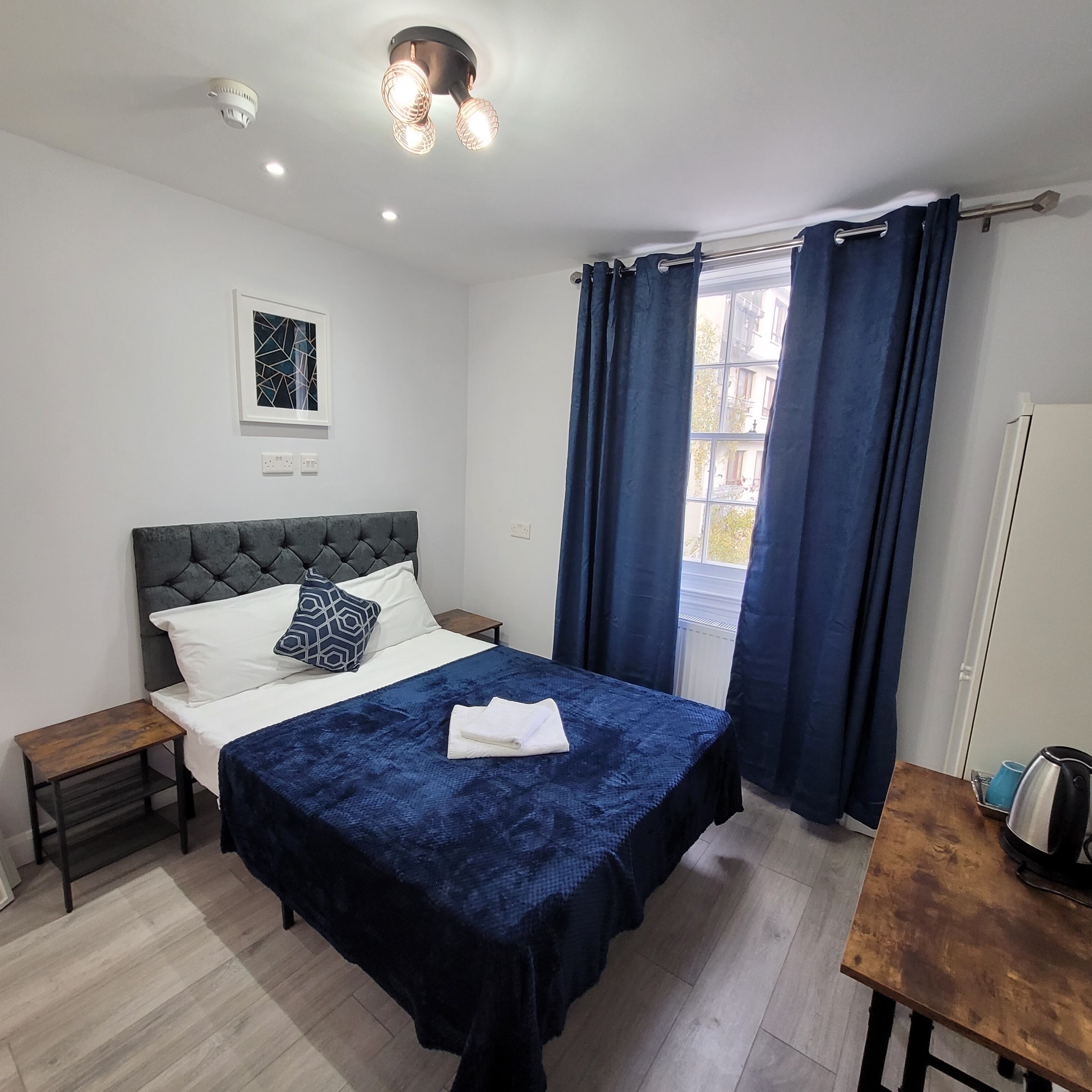 A bedroom with a bed, a nightstand, a small table, a window with curtains, and a ceiling light fixture.