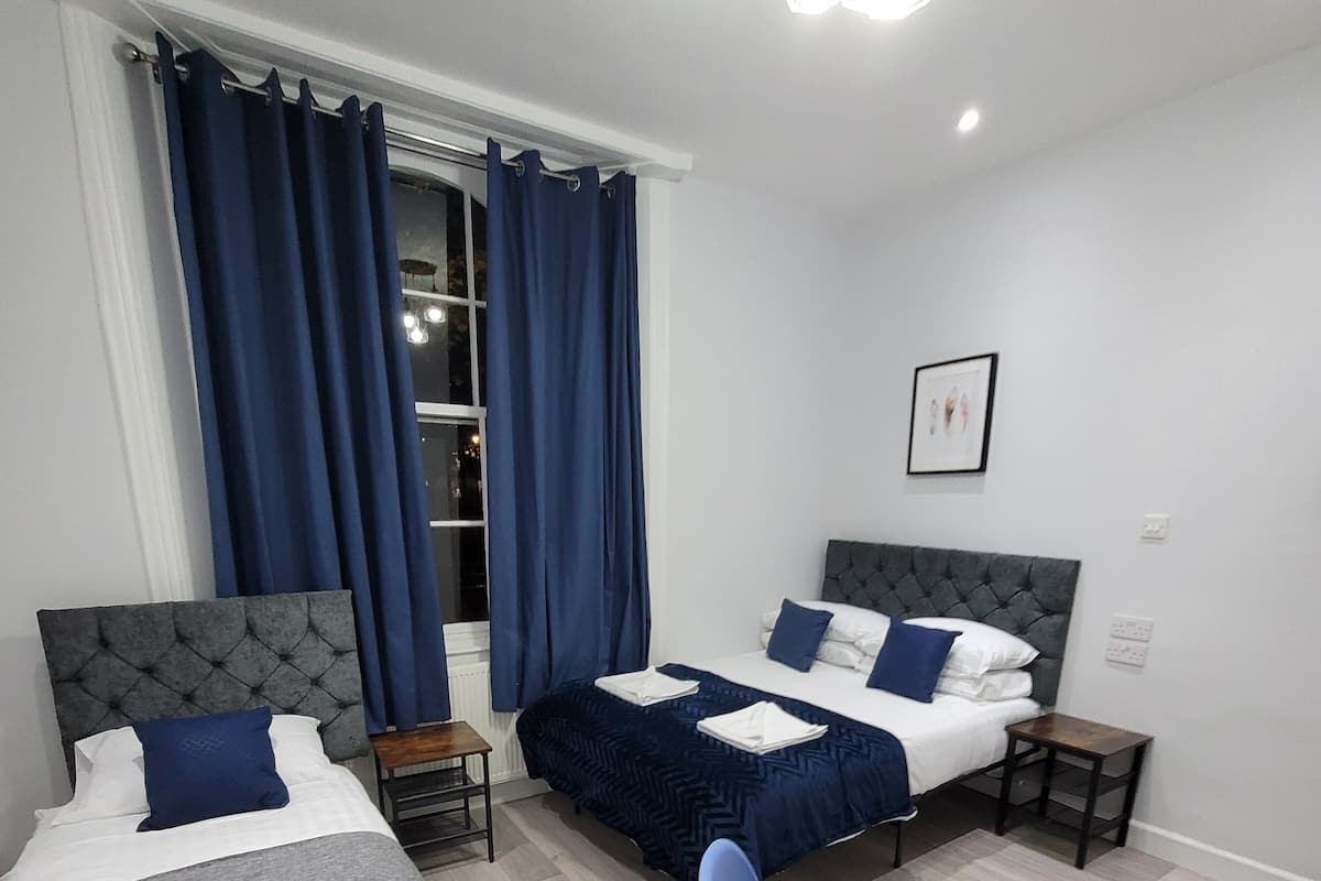 Una habitación de hotel con dos camas, una silla azul, una mesa de madera y un ventanal con cortinas.