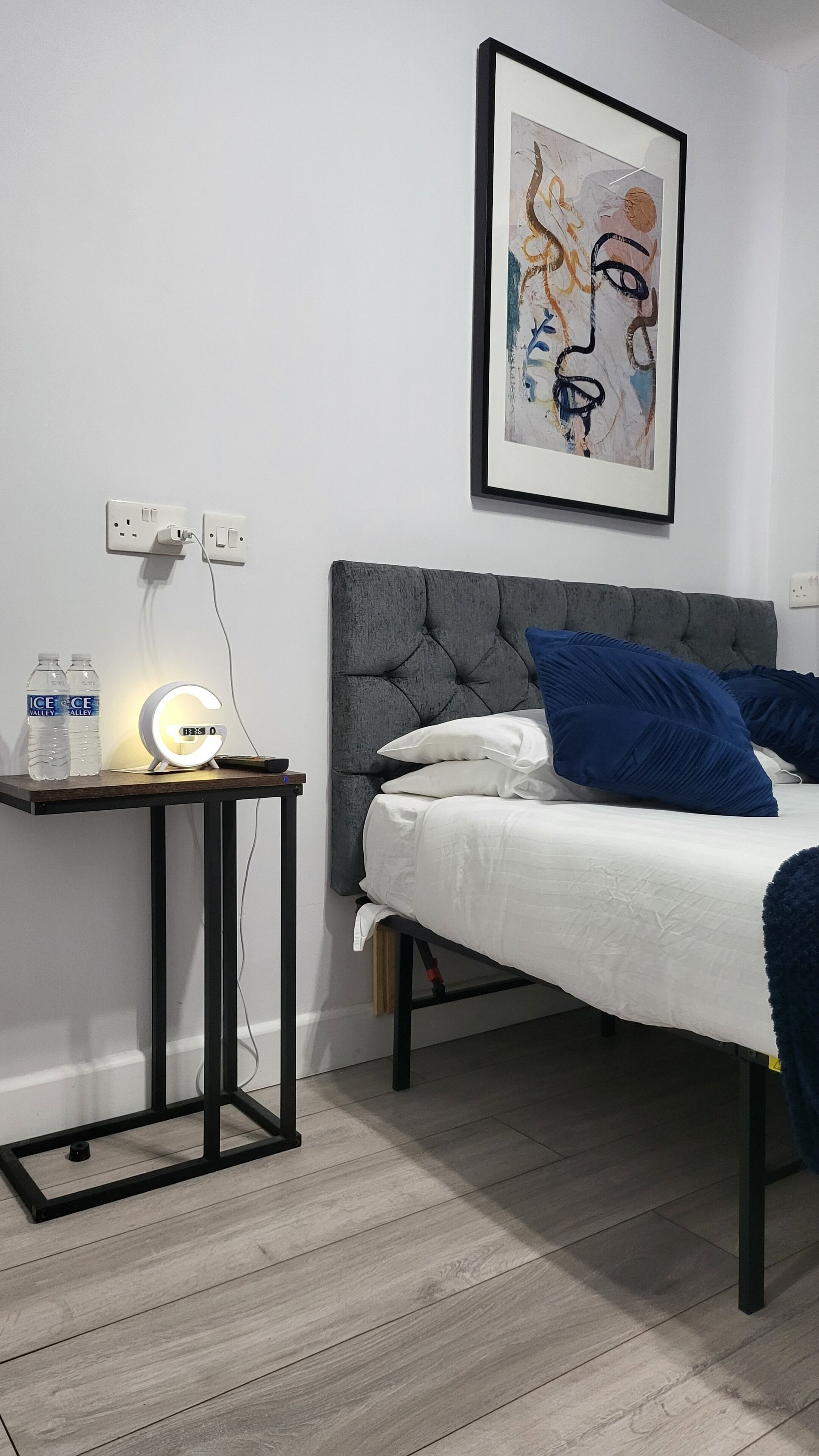 Une chambre à coucher avec un lit, une table de chevet avec une lampe, deux bouteilles d’eau et un tableau abstrait encadré accroché au mur.