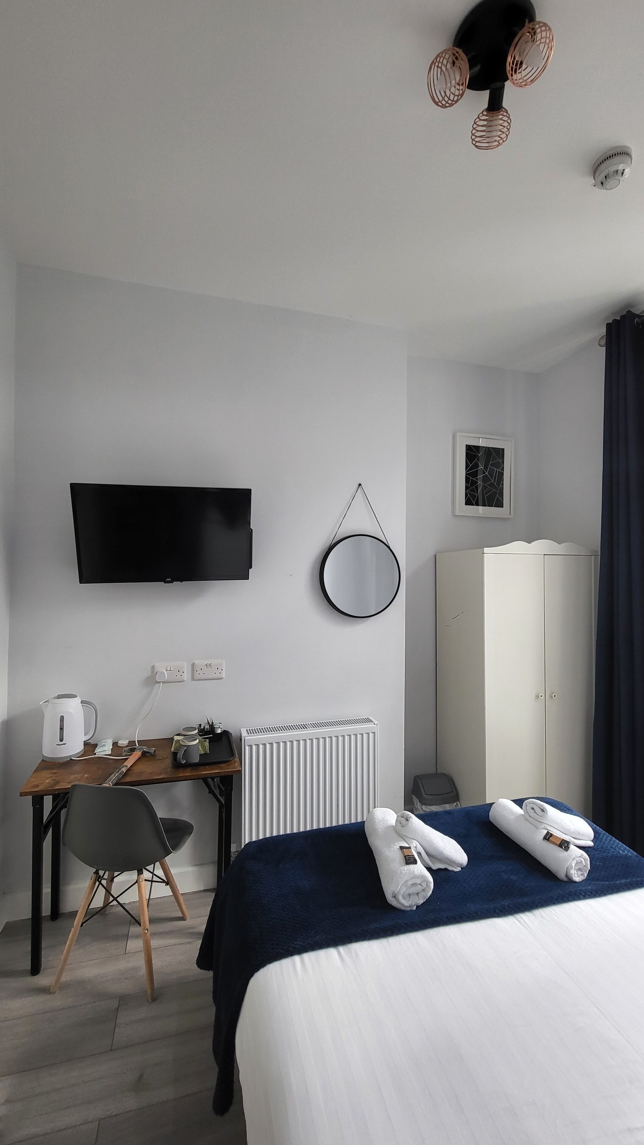 A hotel room with a bed, a desk with a computer, a TV mounted on the wall, a radiator, and a wardrobe.