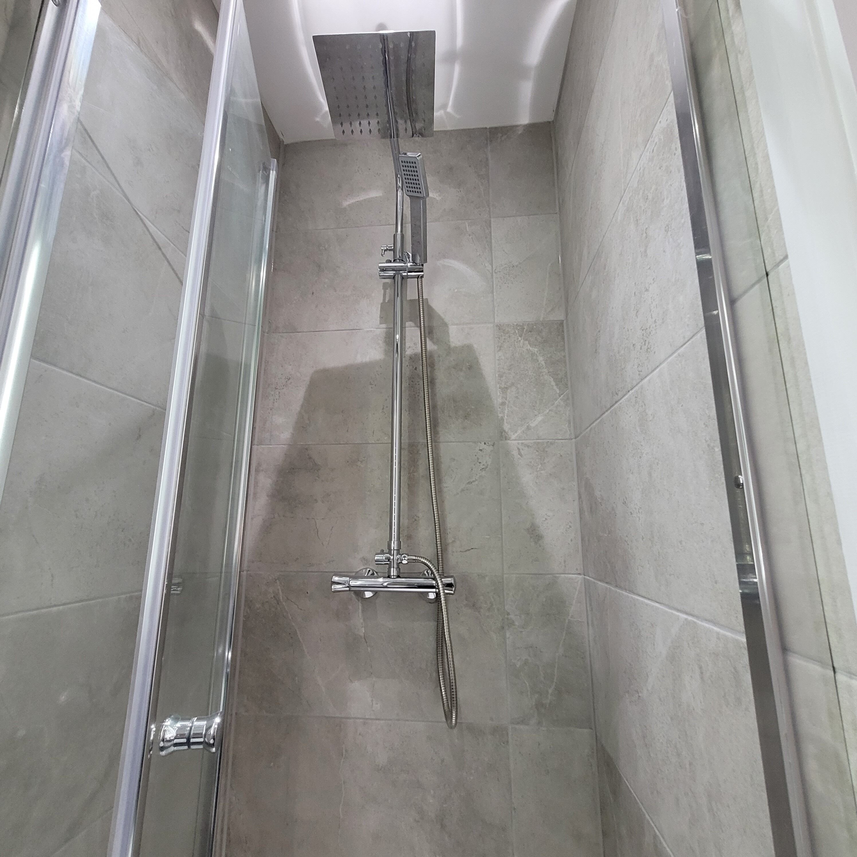 Un box de ducha estrecho con puerta de cristal, cabezal de ducha cromado y suelo de baldosas.