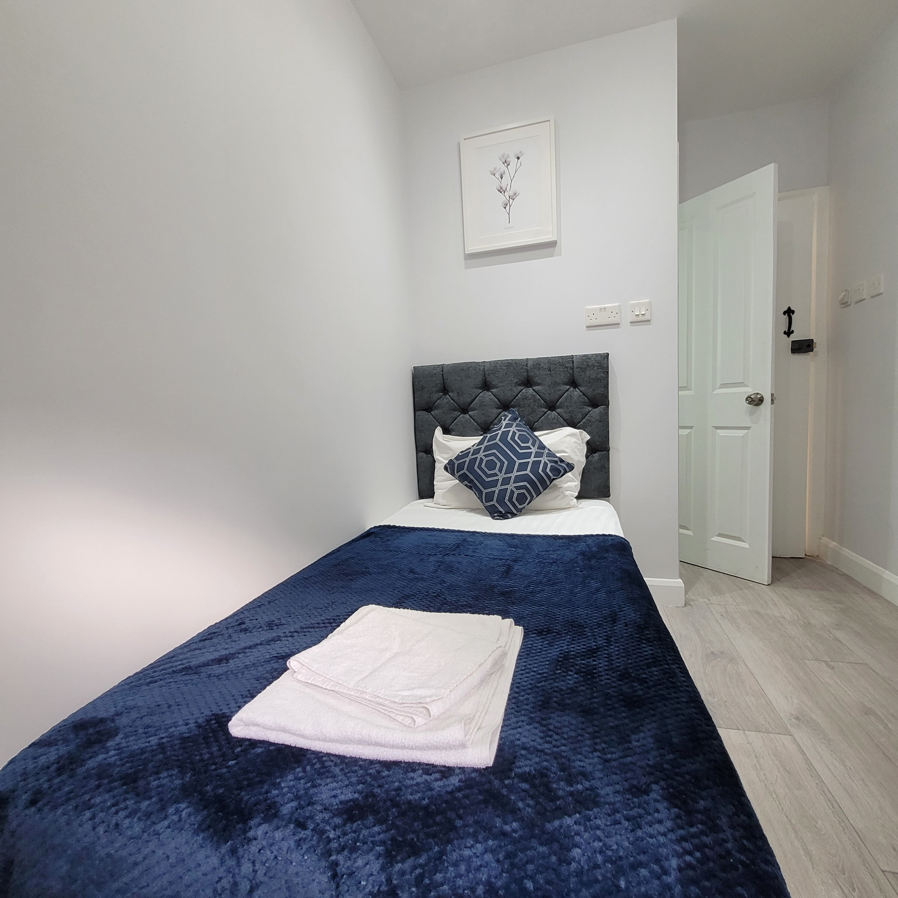 A bedroom with a bed, a blue blanket, a white towel, a grey headboard, and a door.
