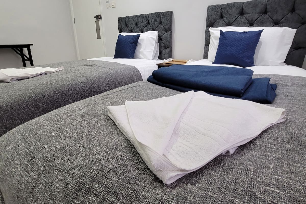 Habitación de hotel con dos camas, cabeceros tapizados en gris, ropa de cama blanca y azul, y una pequeña mesa con una silla.