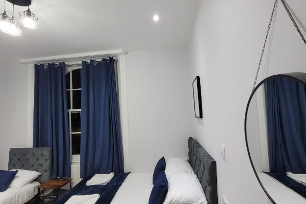 A hotel room with two beds, a round mirror, a chair, and a window with curtains.