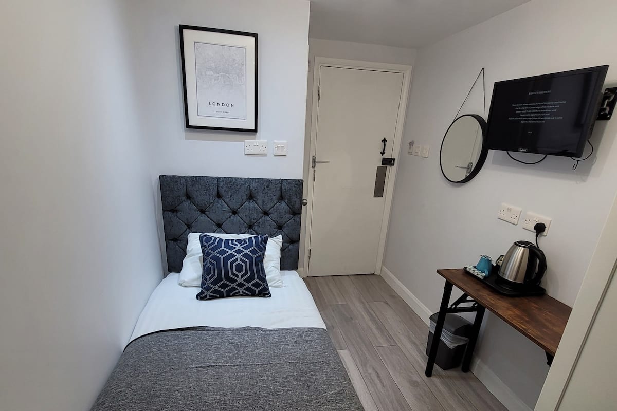 Una habitación de hotel pequeña con una cama, una mesita de noche con una tetera, un televisor y un cuadro en la pared.