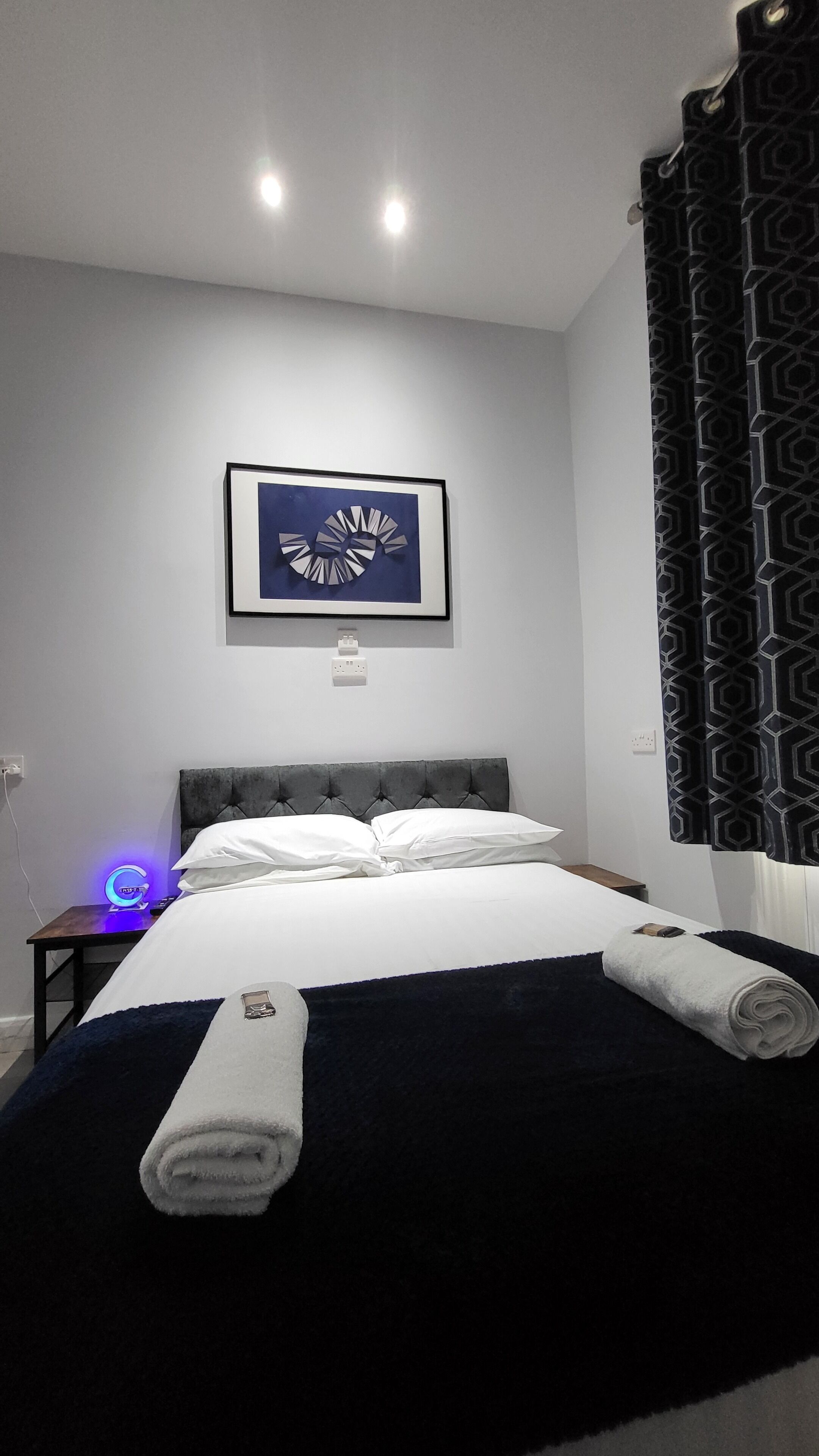 Un lit bien fait, avec du linge de lit blanc, une tête de lit sombre et une œuvre d’art bleue accrochée au mur au-dessus.