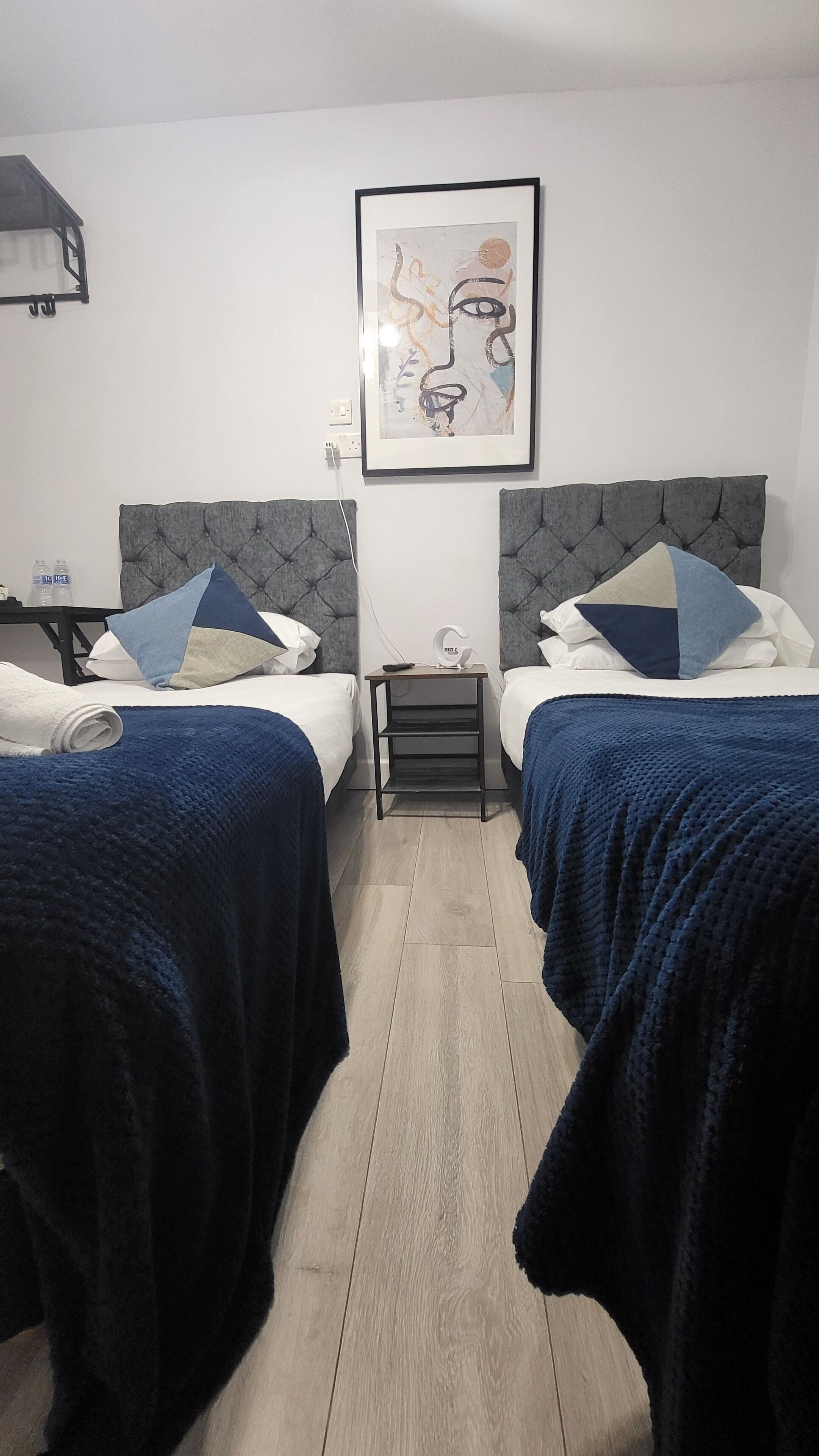 A hotel room with two beds, each with a grey headboard and blue bedding. There is a small bedside table with a lamp, a framed abstract artwork on the wall, and a water bottle on the nightstand.
