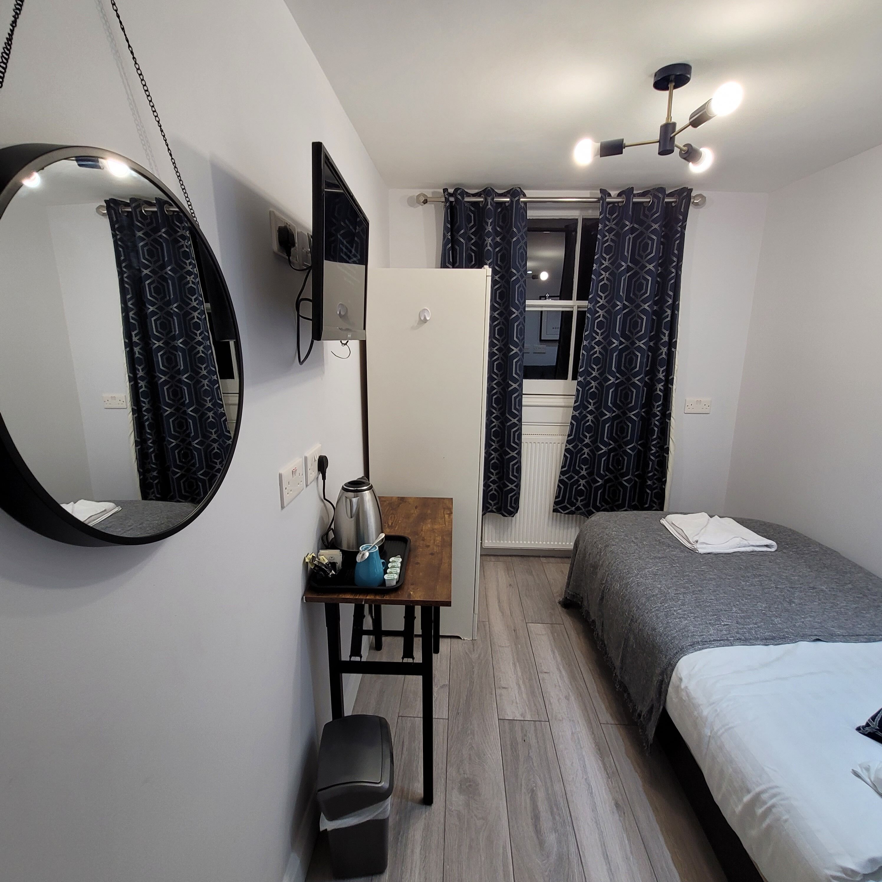 A compact bedroom with a bed, a desk with a kettle, a mirror, and a ceiling fan.