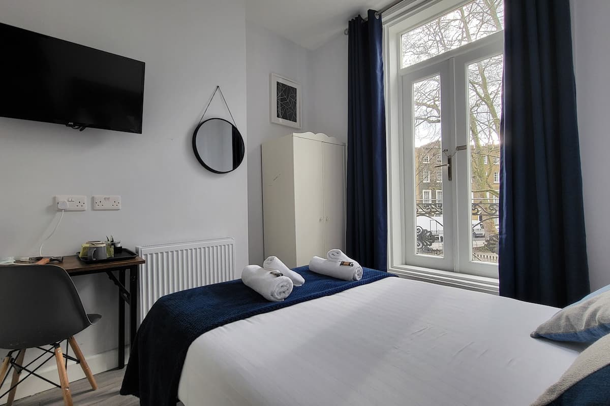 Una habitación de hotel con una cama, un escritorio con hervidor, una silla, un armario, un televisor y una ventana con cortinas.