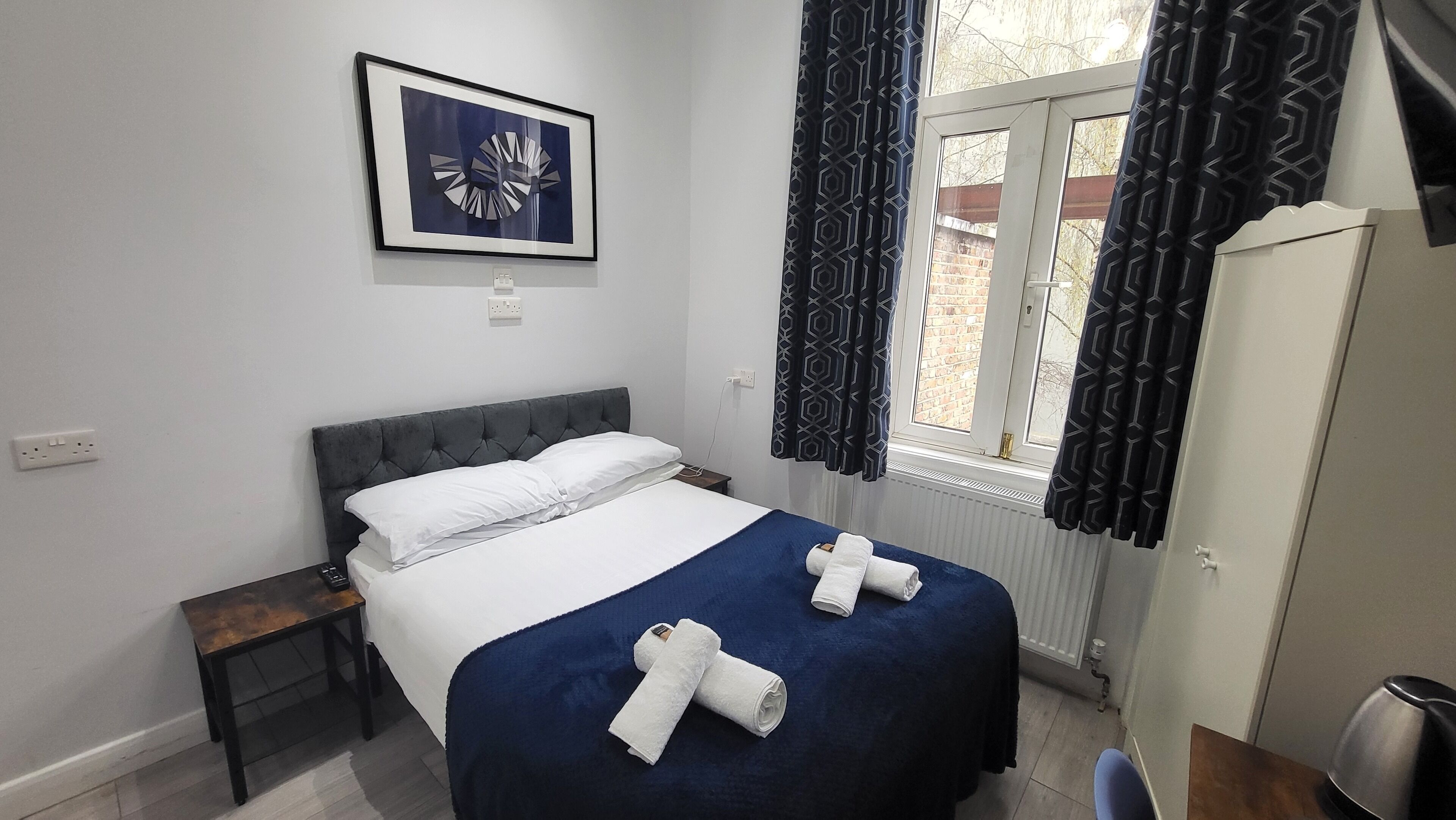A neatly made bed with white linens and two rolled towels, a bedside table with a lamp, a framed abstract artwork above the bed, and a window with patterned curtains.