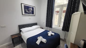 Una cama bien hecha con sábanas blancas y dos toallas enrolladas, una mesita de noche con una lámpara, una obra de arte abstracta enmarcada sobre la cama y una ventana con cortinas estampadas.