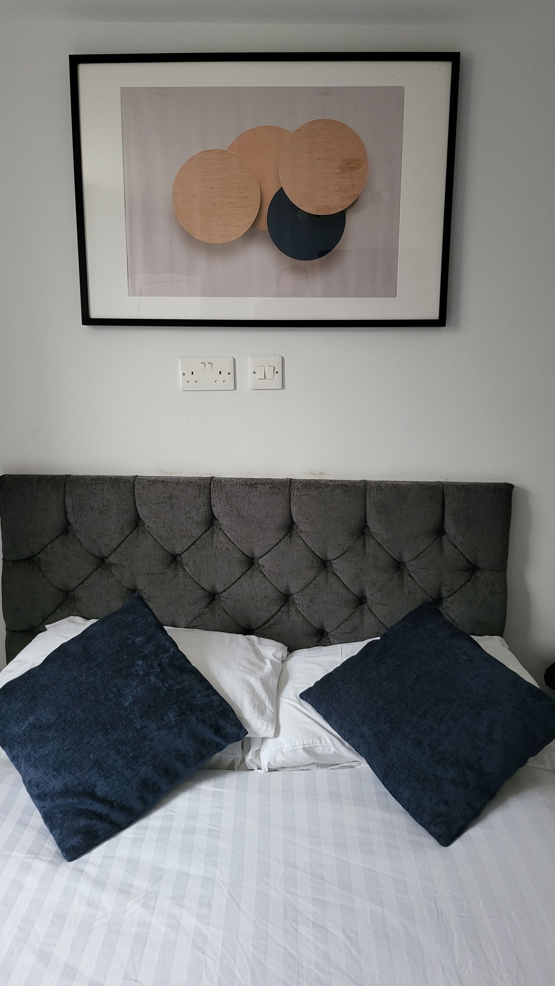 Ein ordentlich bezogenes Bett mit einer gesteppten Kopfteil, zwei dunkelblauen Kissen und einem gerahmten Kunstwerk darüber.