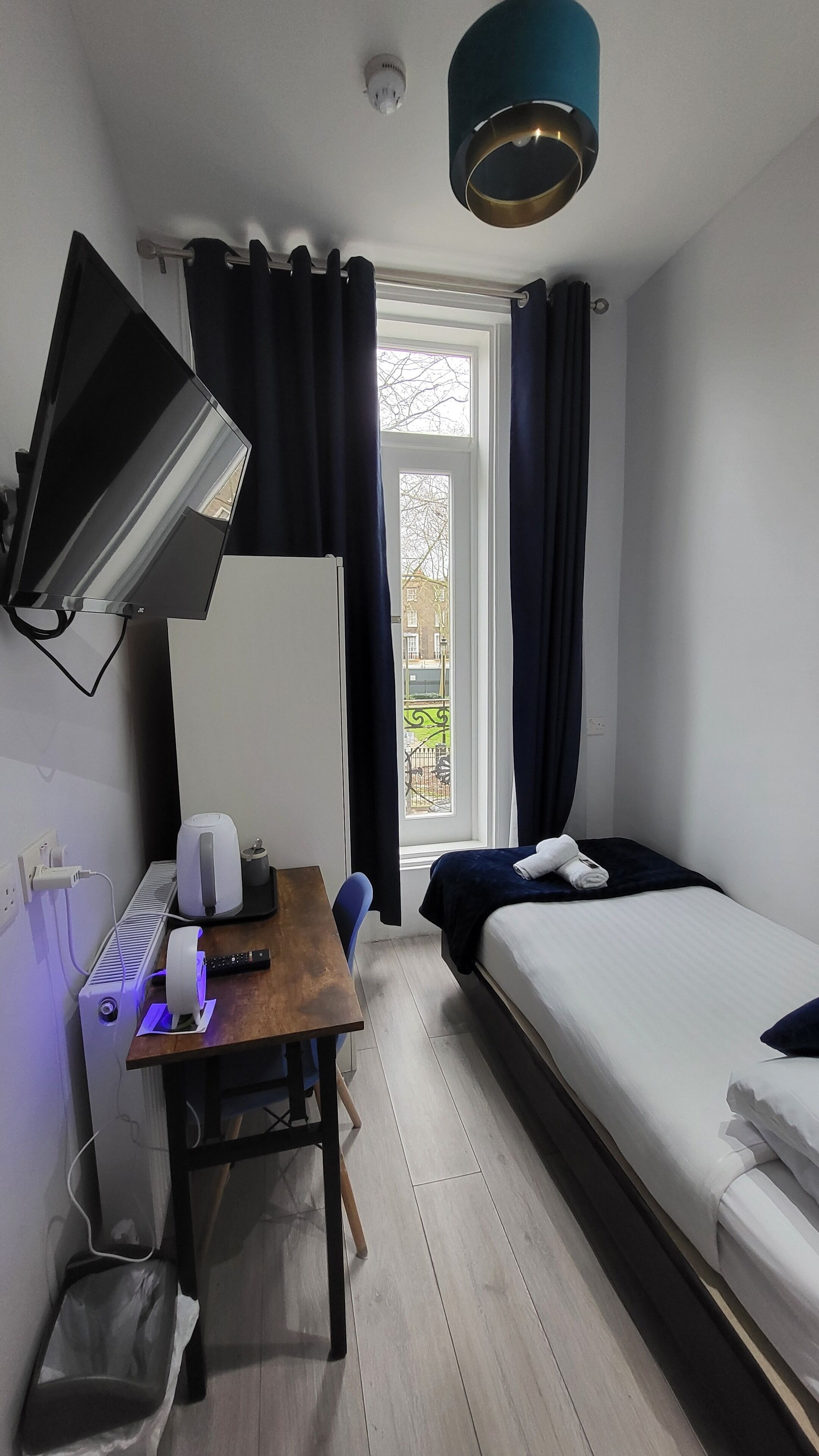 Une chambre d’hôtel compacte comprenant un lit, un bureau avec un ordinateur, un téléviseur à écran plat fixé au mur, une fenêtre avec des rideaux et un luminaire au plafond.