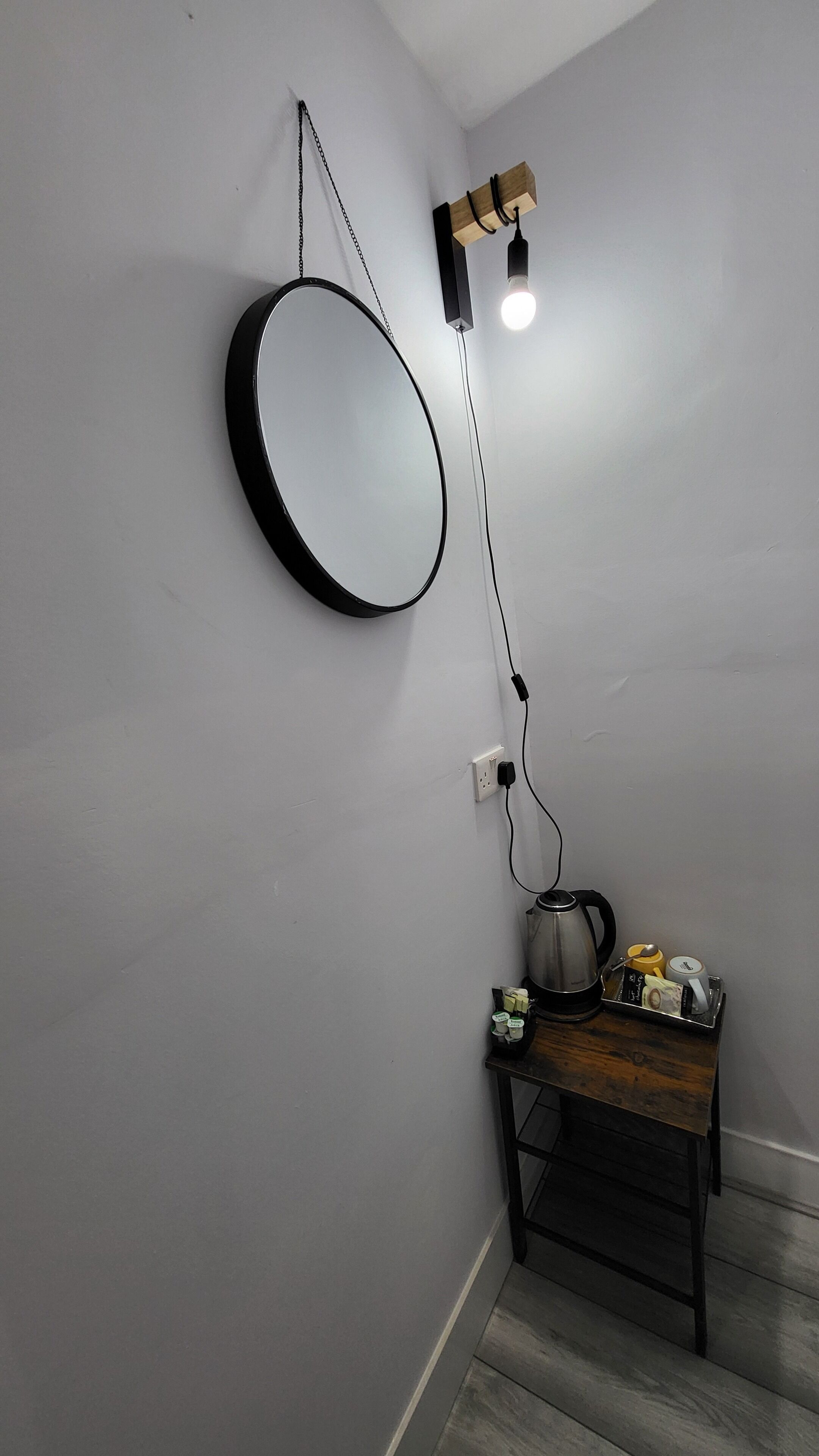 A room with a round mirror on the wall, a table with a kettle, cups, and a tray, and a hanging light fixture.